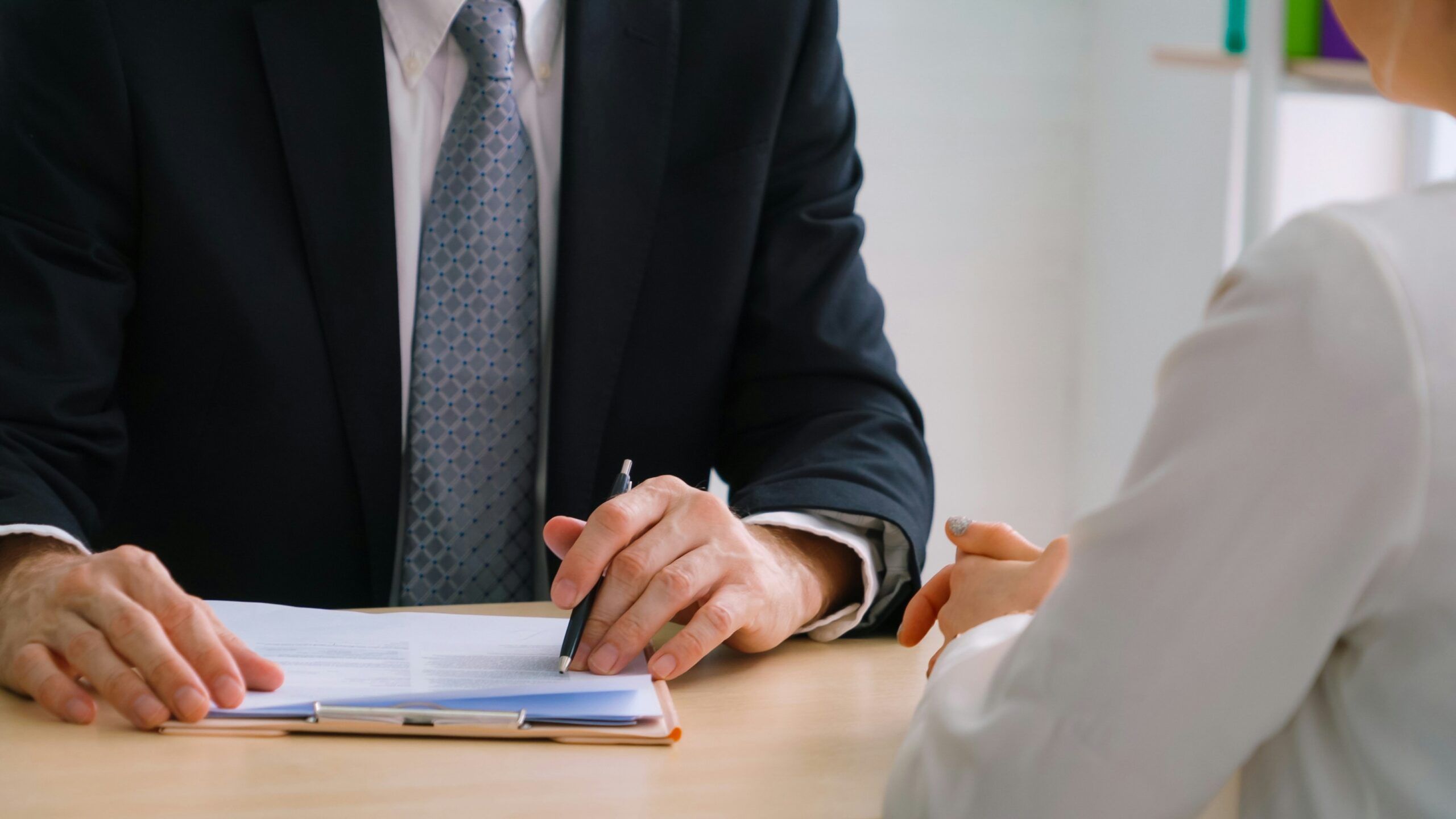 Close-up view of a professional meeting at a desk, showing a person in a dark business suit and patterned tie holding a pen and pointing to a document clipped to a clipboard on a light wooden table. Across from them, another person in a light-colored jacket sits with hands clasped, listening. The focus is on the hands, pen, and paperwork, with faces mostly out of frame, conveying a formal discussion or document review in an office setting.