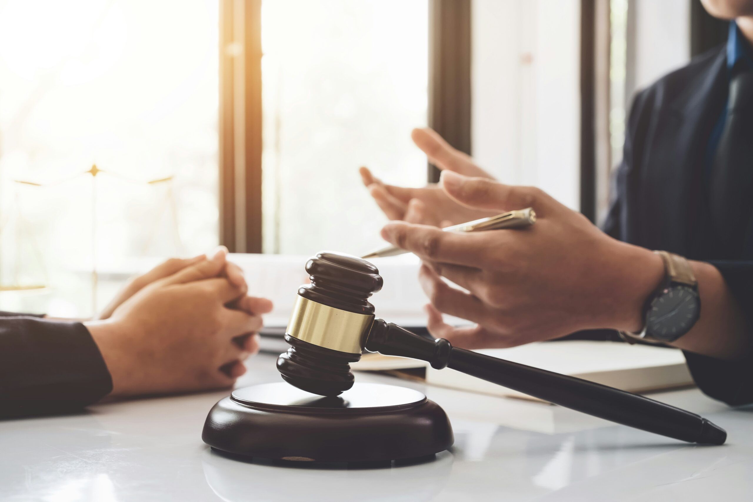 Wooden judge’s gavel resting on a round sound block on a white desk, positioned between two people seated across from each other. One person gestures with a pen while speaking, wearing a dark jacket and wristwatch, while the other listens with hands clasped. Bright daylight streams through a window in the background, suggesting a legal consultation or discussion in an office setting.