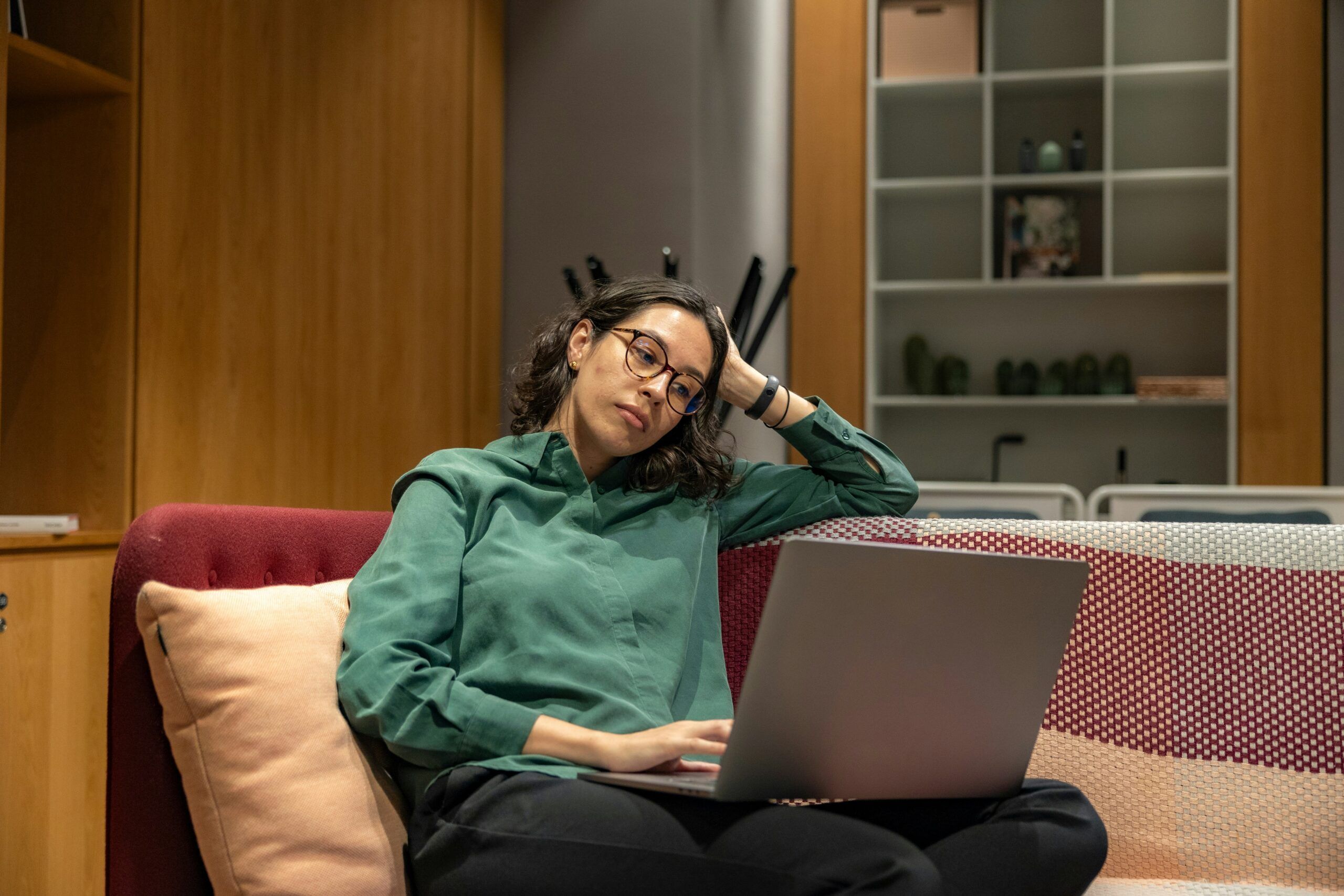 Woman wearing glasses and a green blouse sitting on a sofa with a laptop on her lap, leaning her head on one hand with a tired or contemplative expression, surrounded by a modern interior with wooden cabinetry, shelves, and soft ambient lighting suggesting a home office or lounge workspace.