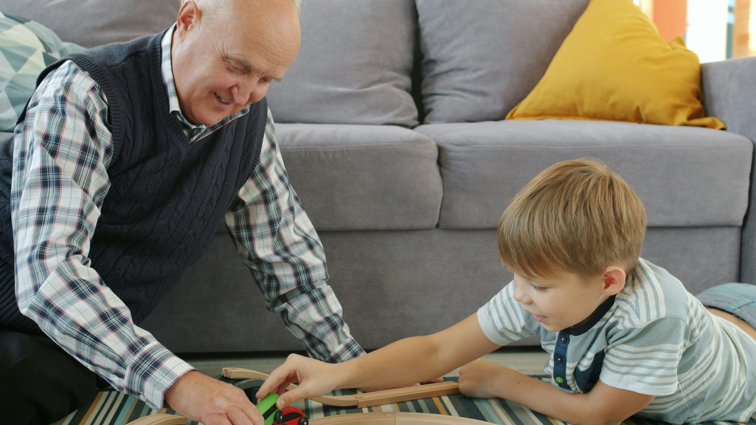 Elderly man and young child sitting on the floor in a living room, smiling as they play together with a wooden toy train set on a rug in front of a gray sofa with colorful cushions.