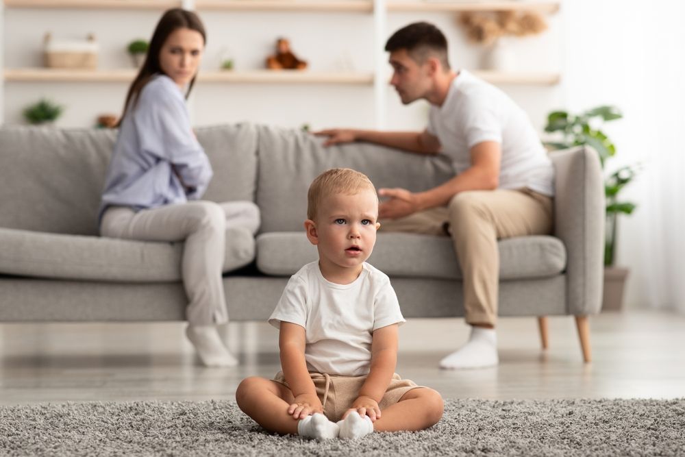 A baby sitting on the floor with a man and woman arguing in the background.