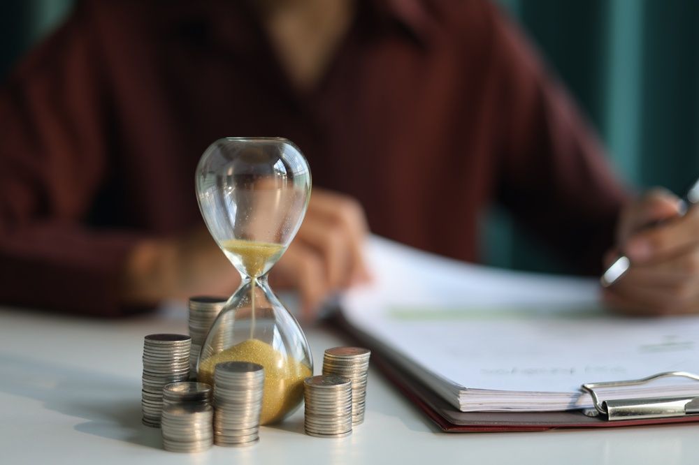 Person writing on paper, with an hourglass and coins beside him, symbolizing time management and financial planning.