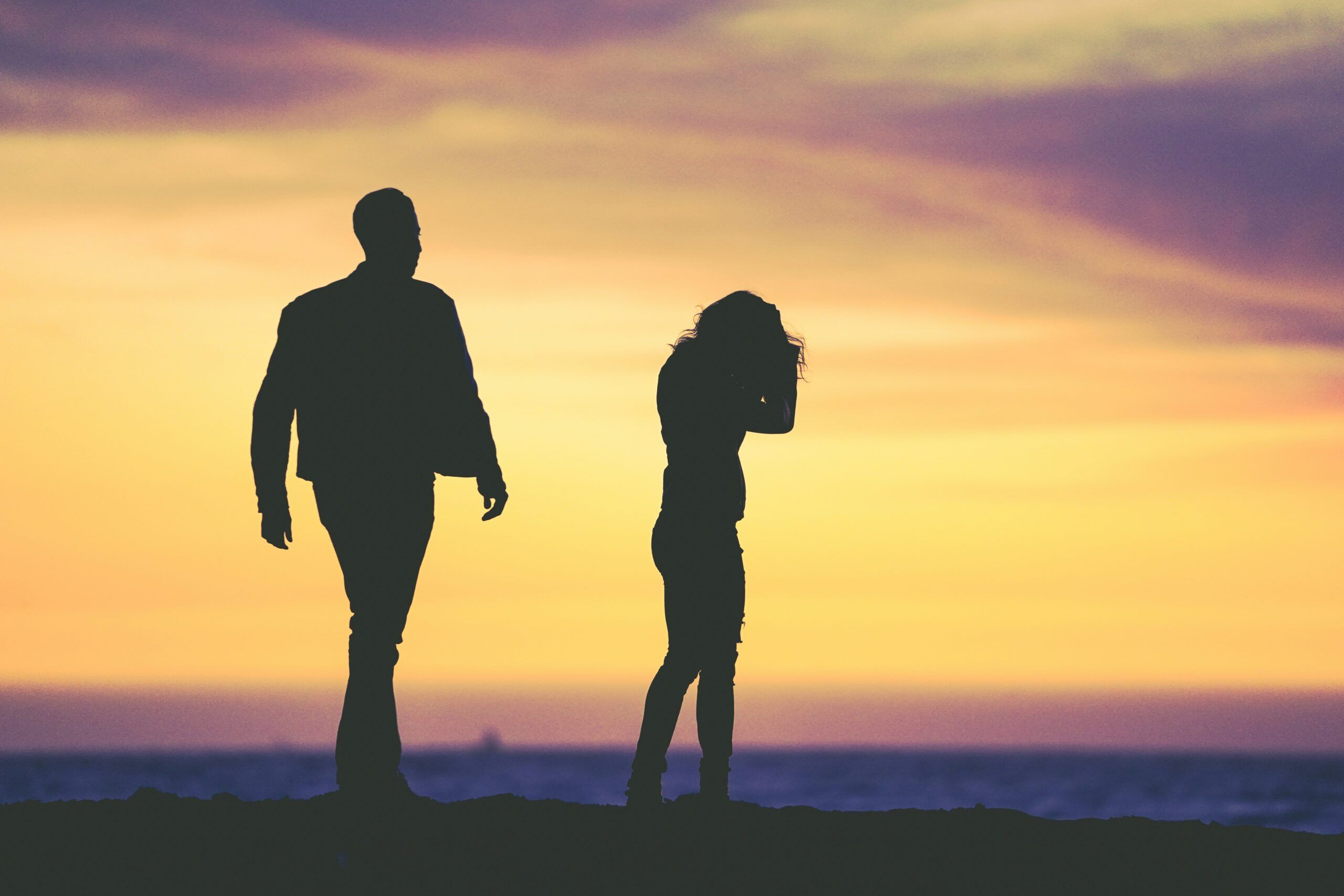 Silhouettes of two people standing apart on a shoreline at sunset, one figure walking away while the other stands turned inward with head lowered and hand near the face, set against a glowing sky of yellow, orange, and purple hues above the dark horizon and calm sea, conveying distance and emotional separation.