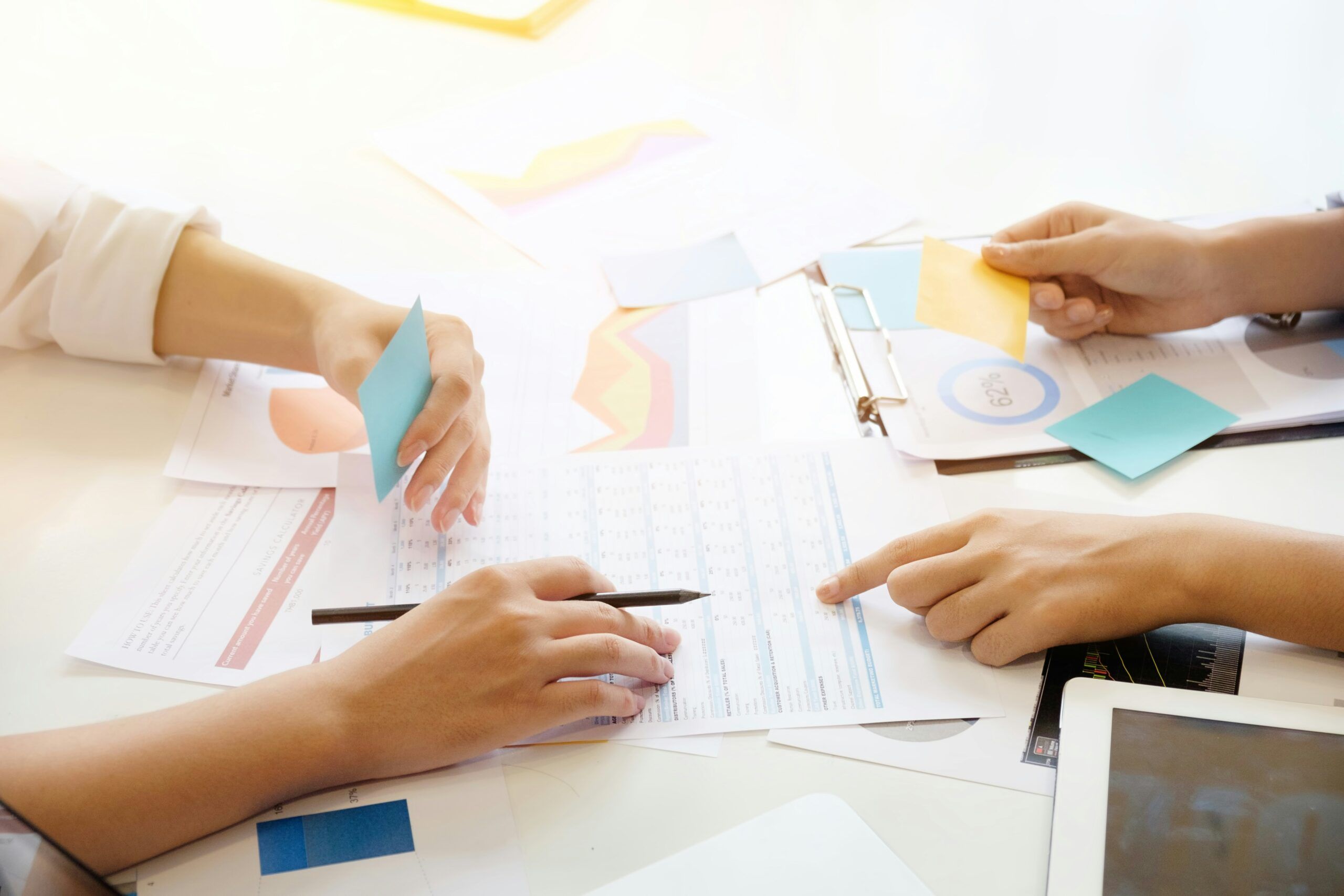 Several people’s hands gathered around a desk covered with printed charts, graphs, and forms, one hand holding a pen over a document, others pointing at specific rows and sections, with colorful sticky notes being held and placed on the papers, suggesting an active discussion or collaborative review of data during a meeting.