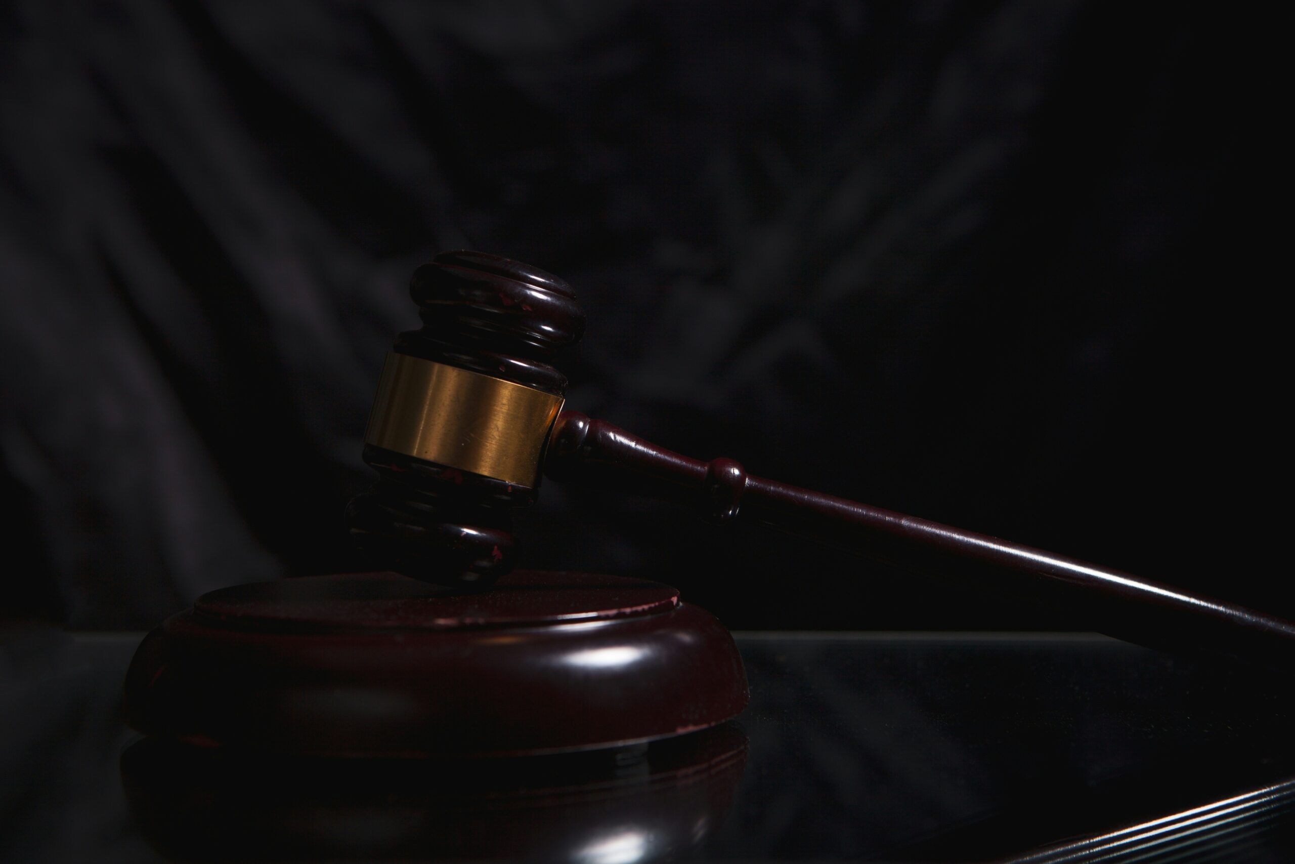 Dark, dramatic close-up of a wooden judge’s gavel with a brass band resting at an angle on its round sound block, positioned on a glossy reflective surface, with soft directional lighting and a deep black background that emphasizes the gavel’s polished texture, curved handle, and the formal, authoritative atmosphere of a courtroom setting.