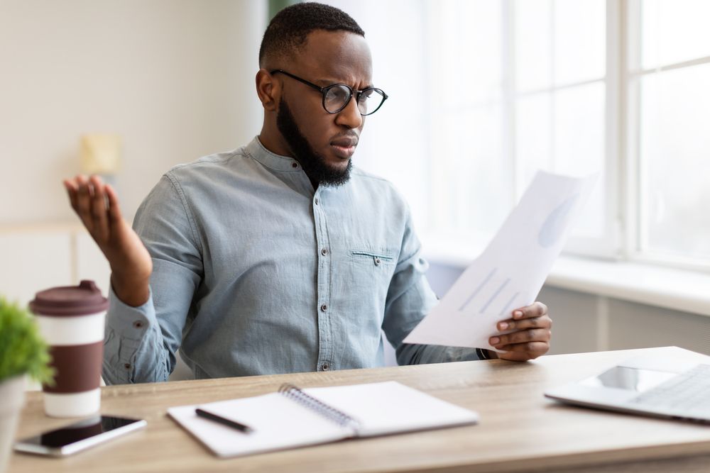 A man with glasses and a beard sits at a desk, looking at the paper