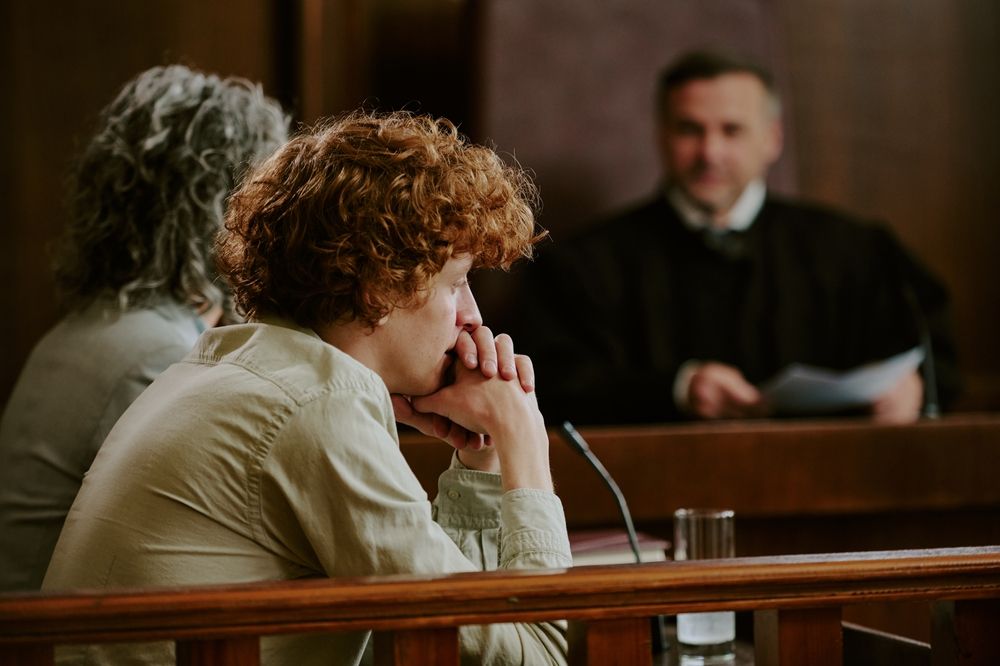 Caucasian teenager sitting at witness stand in courtroom, resting chin on clasped hands, listening attentively while judge in robe observing from bench in background