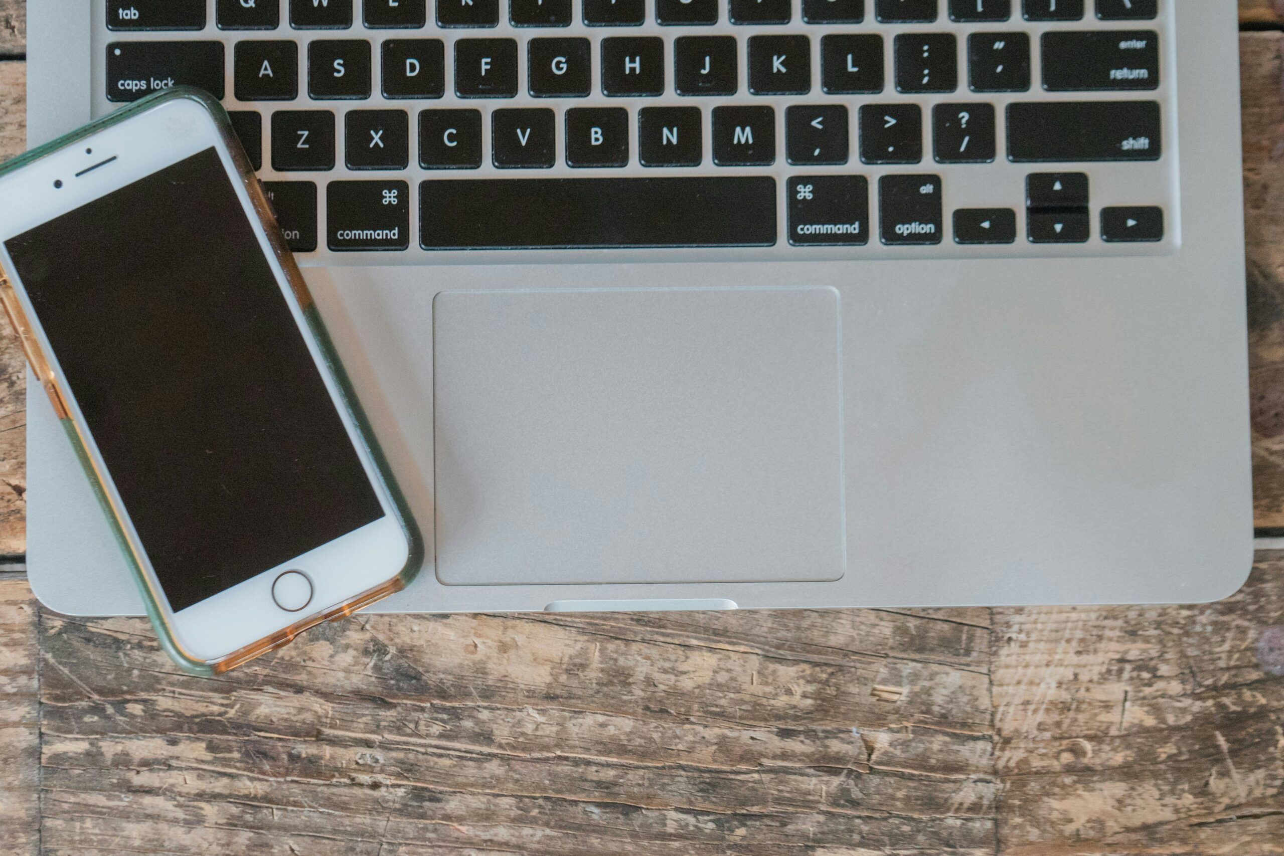 an iPhone rests on the keyboard of a Mac book, which is on a wooden surface
