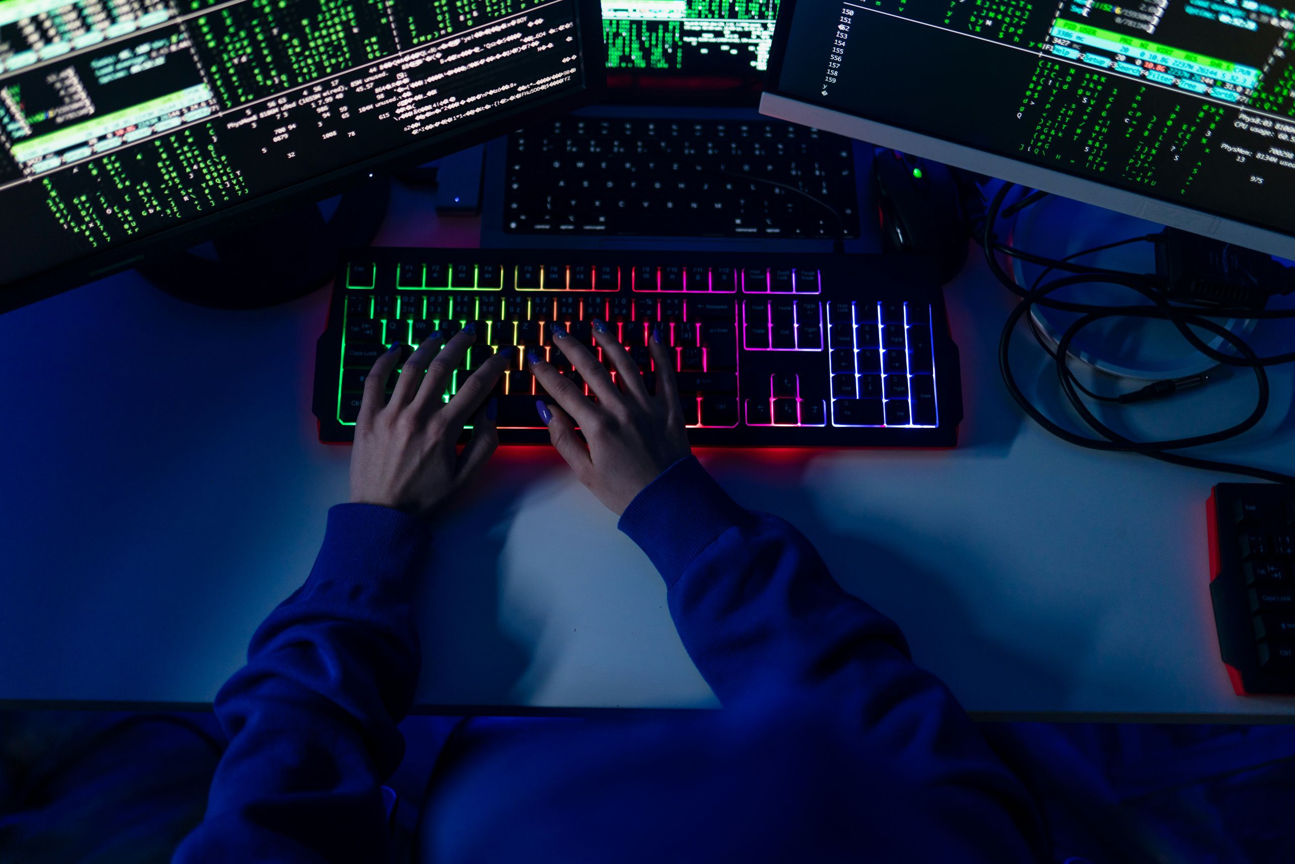 
                    A dimly lit workspace shows a person typing on a brightly illuminated RGB mechanical keyboard, surrounded by multiple monitors filled with lines of code and matrix-style scrolling data.