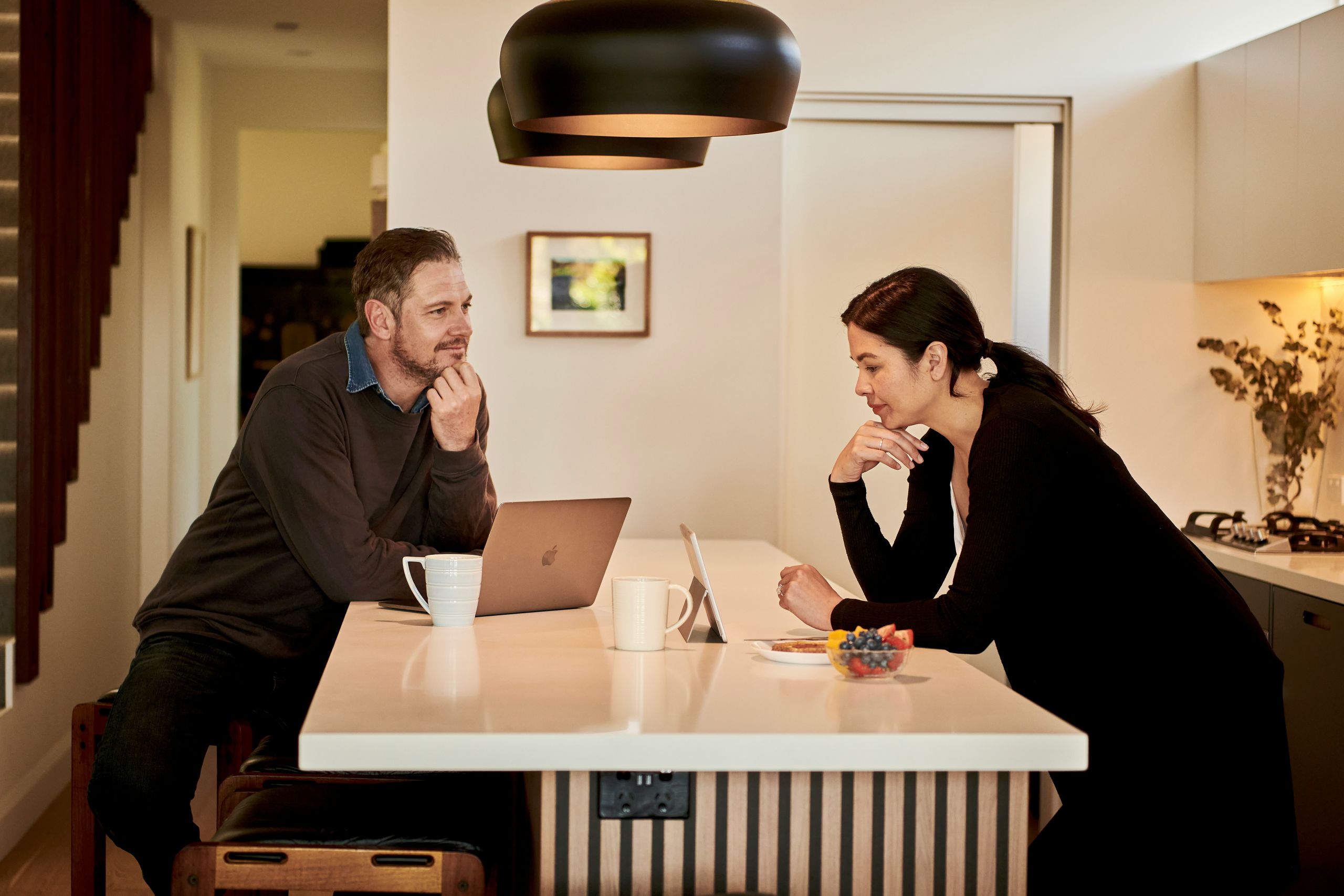 Two adults leaning across a kitchen island in a modern home, facing each other in conversation with open laptops, coffee mugs, and a small plate of fruit on the counter beneath warm pendant lighting.