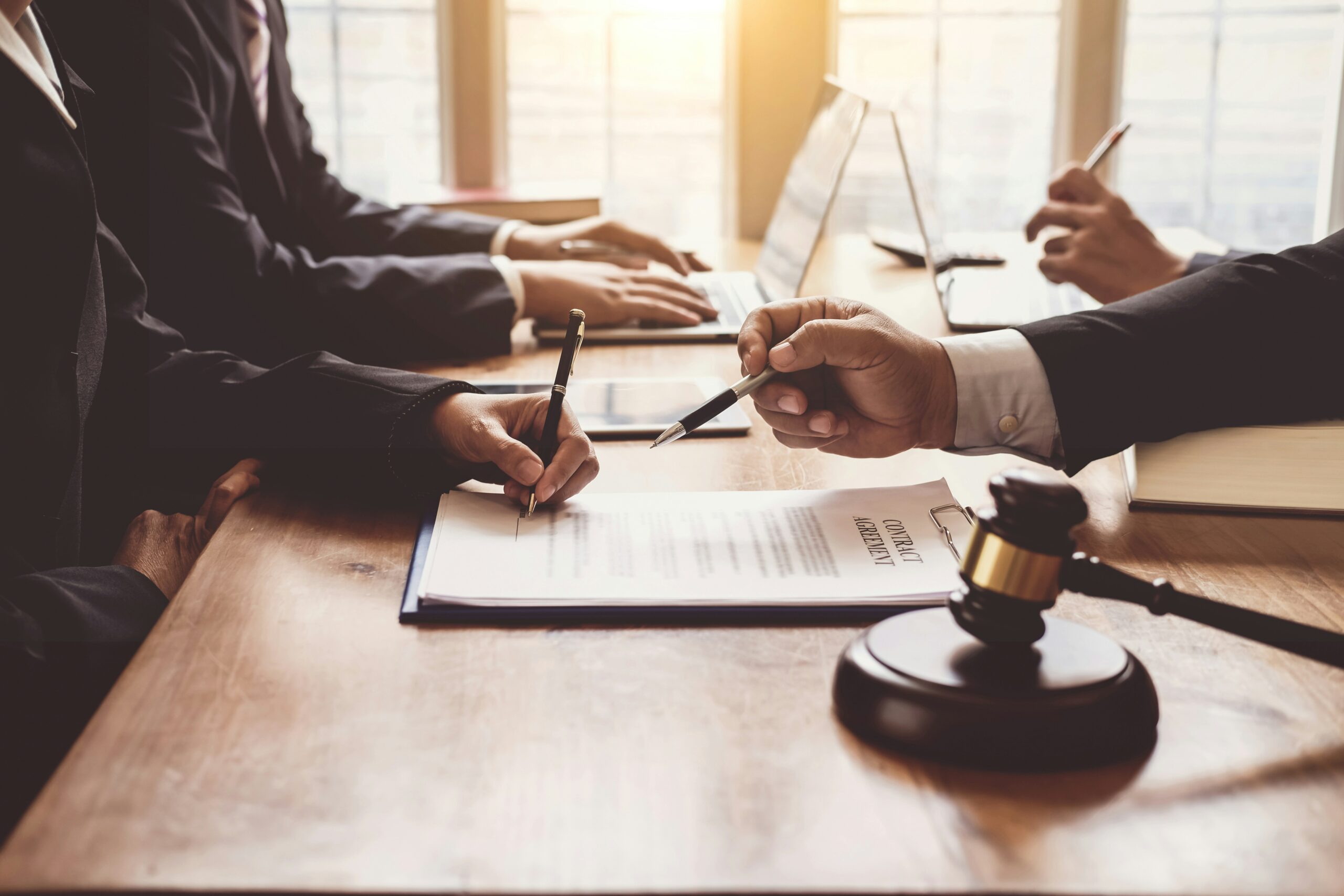 Close-up of several people in business suits seated at a wooden table, signing a printed legal contract with pens, while another person gestures toward the document, with a judge’s gavel resting in the foreground and open laptops visible in the background, suggesting a formal legal or contract negotiation setting.