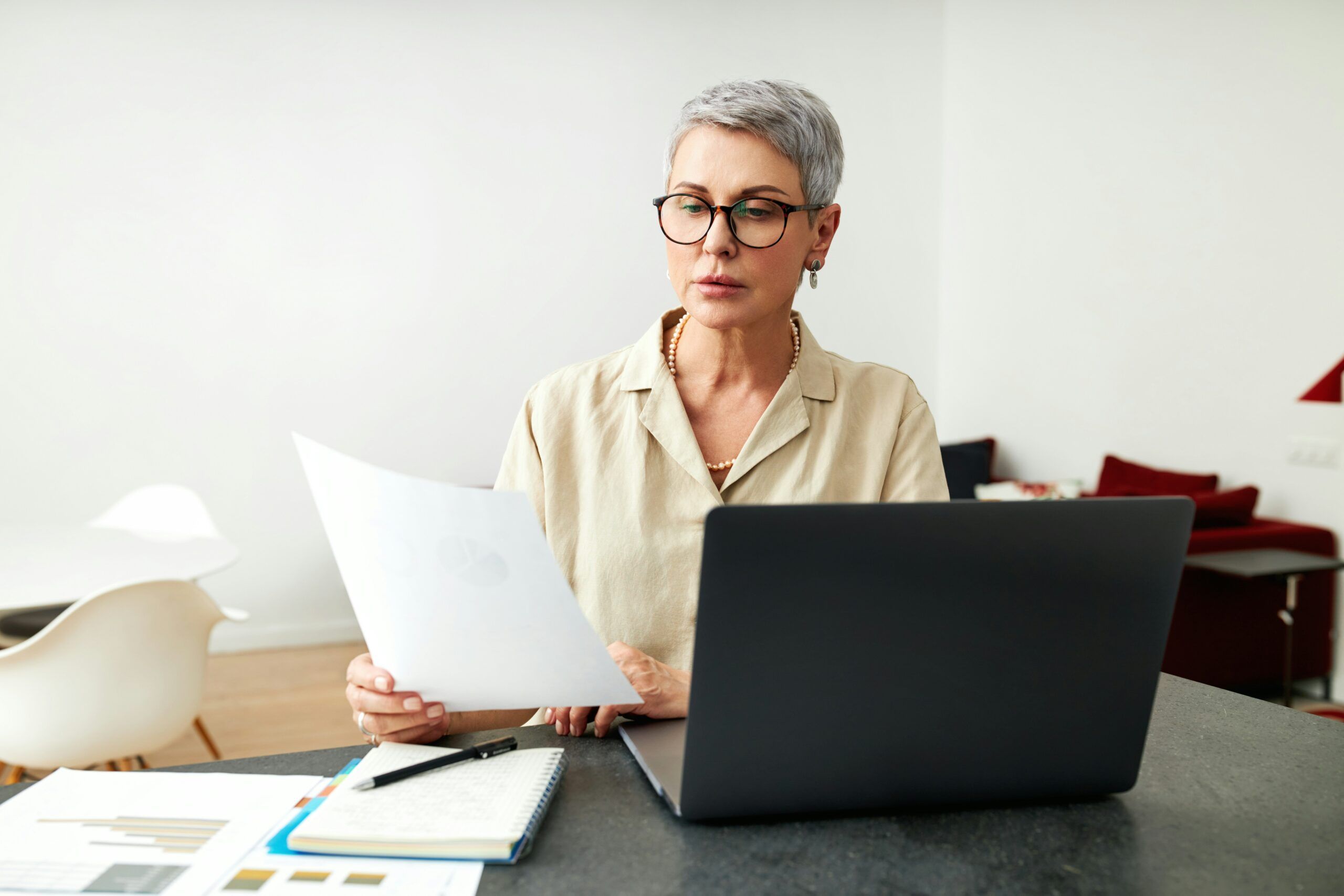 A middle-aged woman with short gray hair and round eyeglasses sits at a table in a bright, minimalist room, holding a single sheet of paper in one hand while looking at an open laptop in front of her. She wears a light beige blouse and a simple necklace, with a focused, thoughtful expression. On the table are a notebook with a pen, printed charts or documents, and the laptop positioned slightly to her right. The background is softly lit and uncluttered, with light-colored walls, modern furniture, and muted red cushions, creating a calm, professional atmosphere.