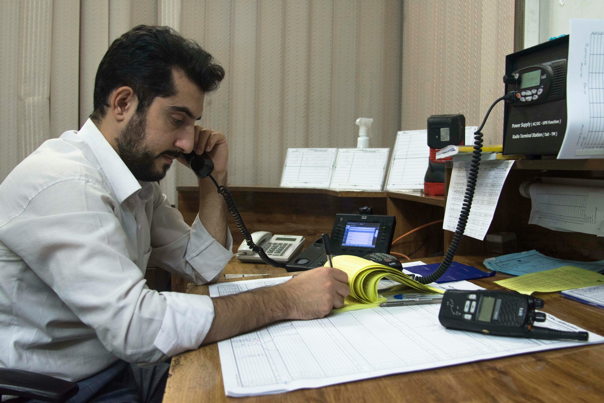 A man with dark hair and a short beard sits at a wooden desk in a small office, holding a corded telephone to his ear with one hand while writing notes in a yellow notepad with the other; he is wearing a white button-down shirt and appears focused and serious. The desk is covered with paperwork, a large open log sheet, pens, and a handheld radio, while additional telephones, communication equipment, and pinned paper notes are arranged on shelves and the wall in front of him, creating a busy, work-oriented environment.