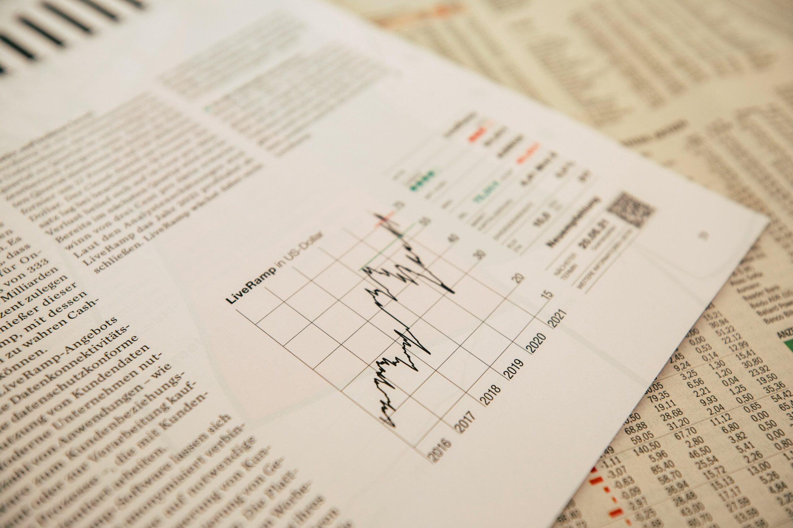 A close-up view of an open printed report or magazine spread showing a financial line chart titled “LiveRamp US Data,” with a fluctuating black line plotted across a grid spanning several years from 2016 to 2021. The chart is surrounded by dense blocks of printed text and numerical tables on adjacent pages, slightly out of focus, suggesting detailed financial or market analysis. The paper lies at an angle on top of other documents, with warm, soft lighting emphasizing the texture of the pages and the contrast between the chart lines, grid markings, and printed data.
