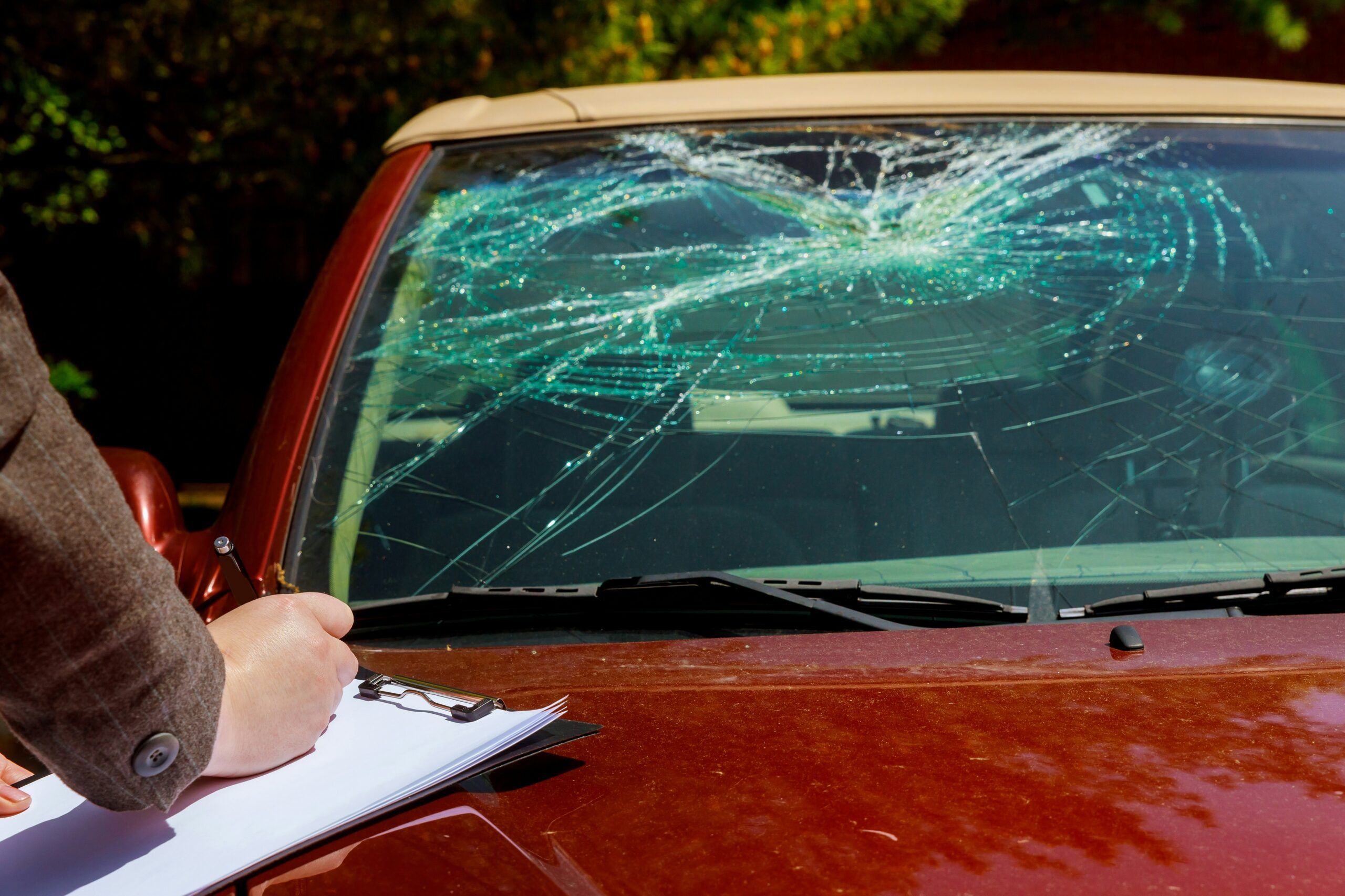 A man writes on a clipboard while examining a car with a shattered windshield.