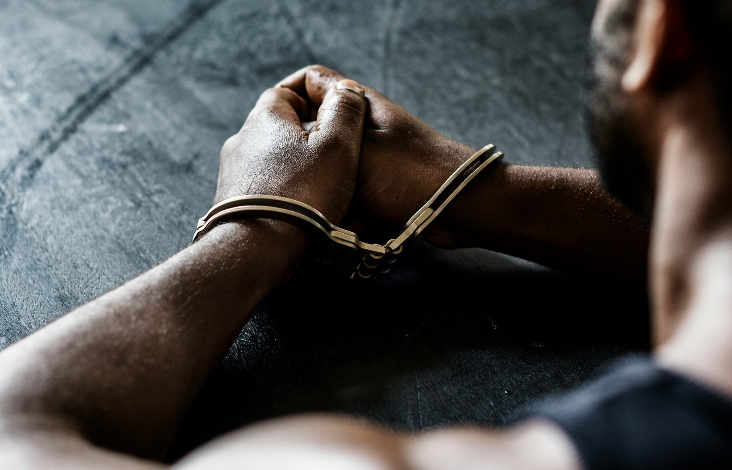 A man in handcuffs sits on a table, appearing calm and composed in a stark, minimalistic setting.