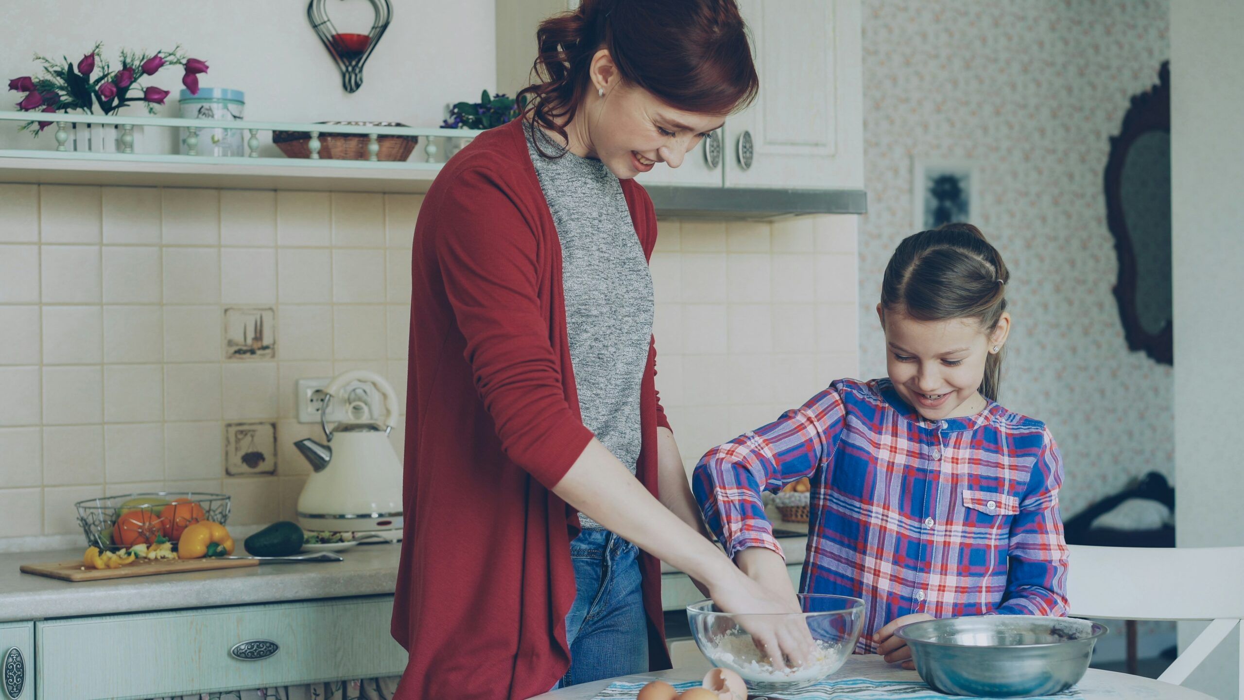 A happy mother and child in a brightly lit kitchen. Their hands are in a clear mixing bowl with flour