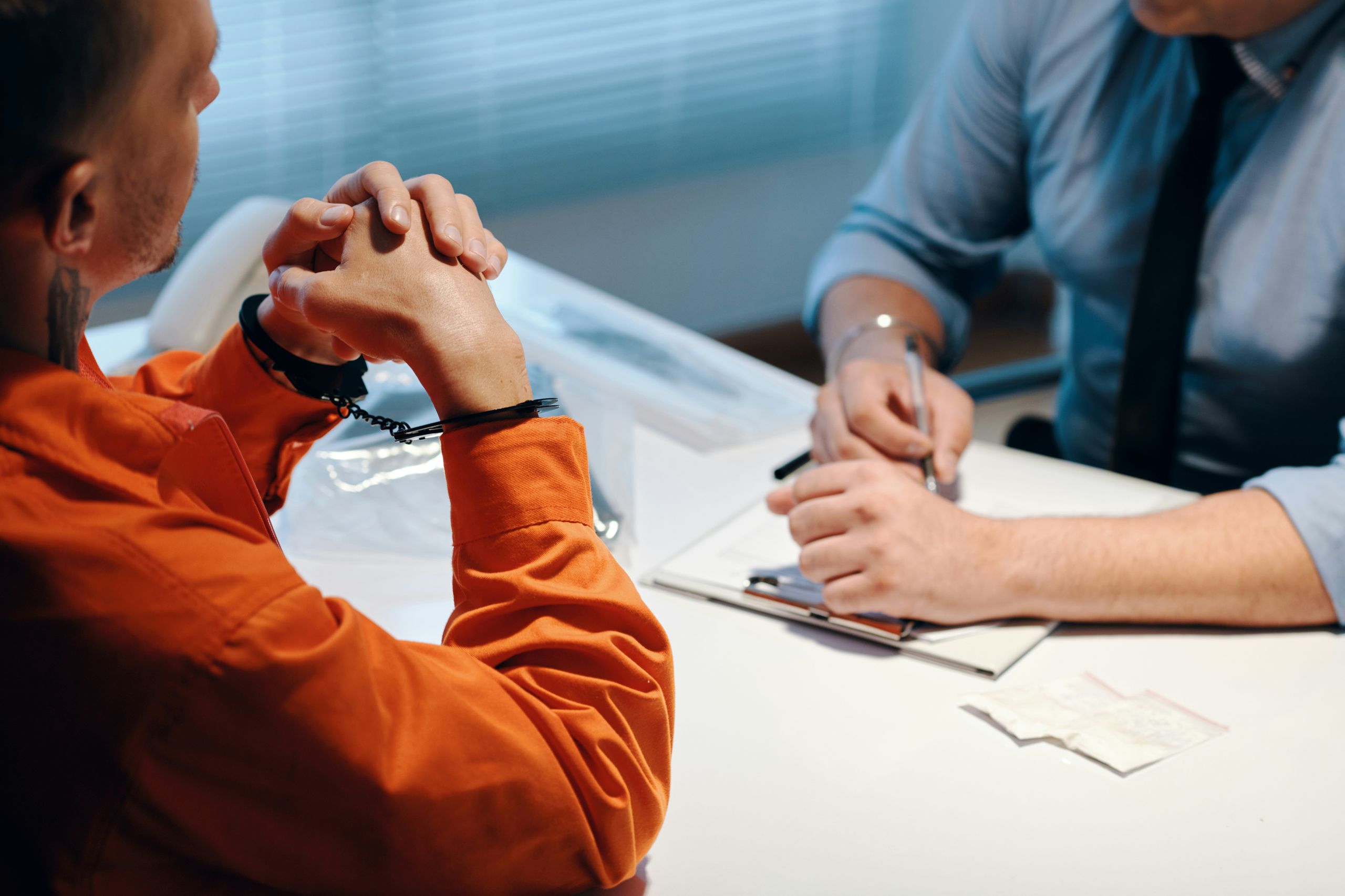 a man in an orange jumpsuit is seated at a table. his elbows are on the table and he is handcuffed. A police officer sits across from him with paperwork.