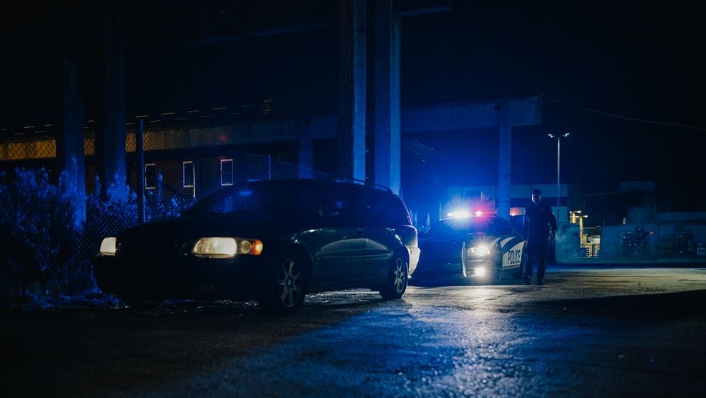 Officer approaching a stopped car at night beside a police vehicle with flashing lights