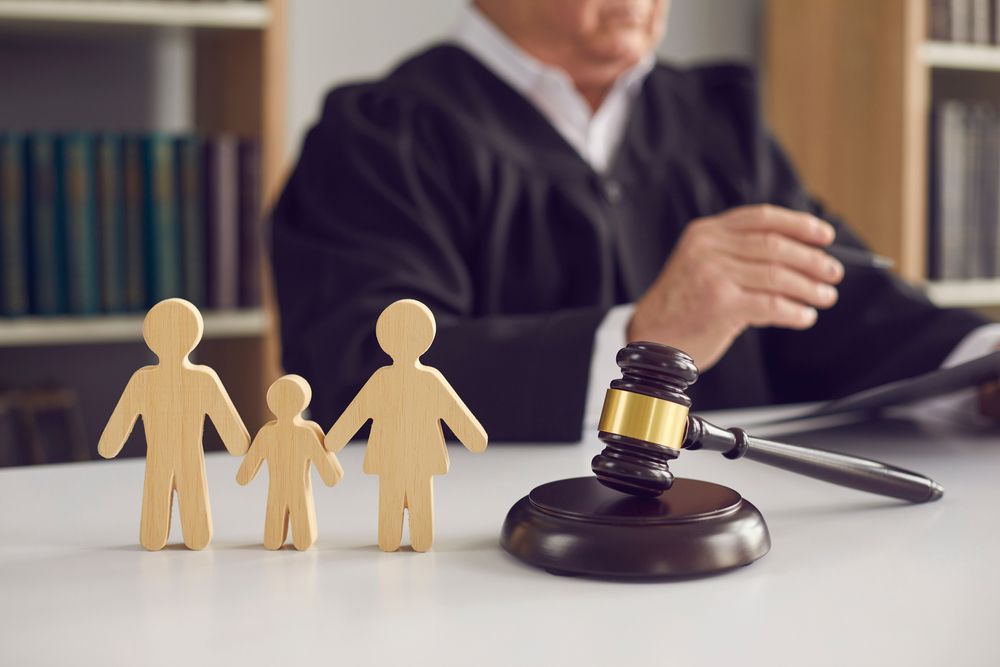 Wooden cutout figures of two adults and a child beside a gavel, with a judge at a desk behind
