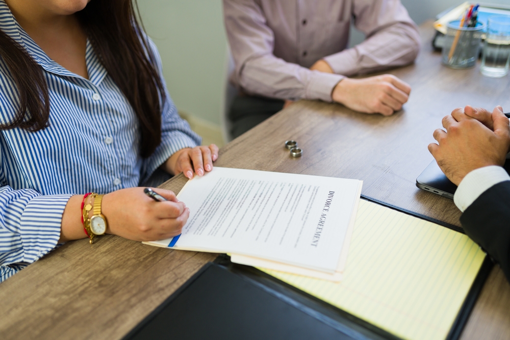 Woman signing a document titled Divorce Agreement at a table with a man and an attorney, with two wedding rings on the table